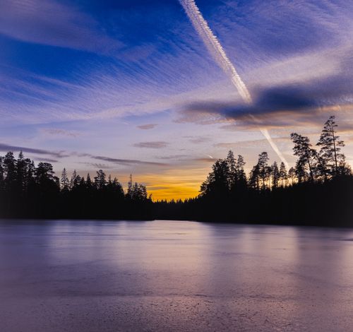 Free A peaceful sunset view over a calm lake with silhouetted trees and colorful sky. Stock Photo