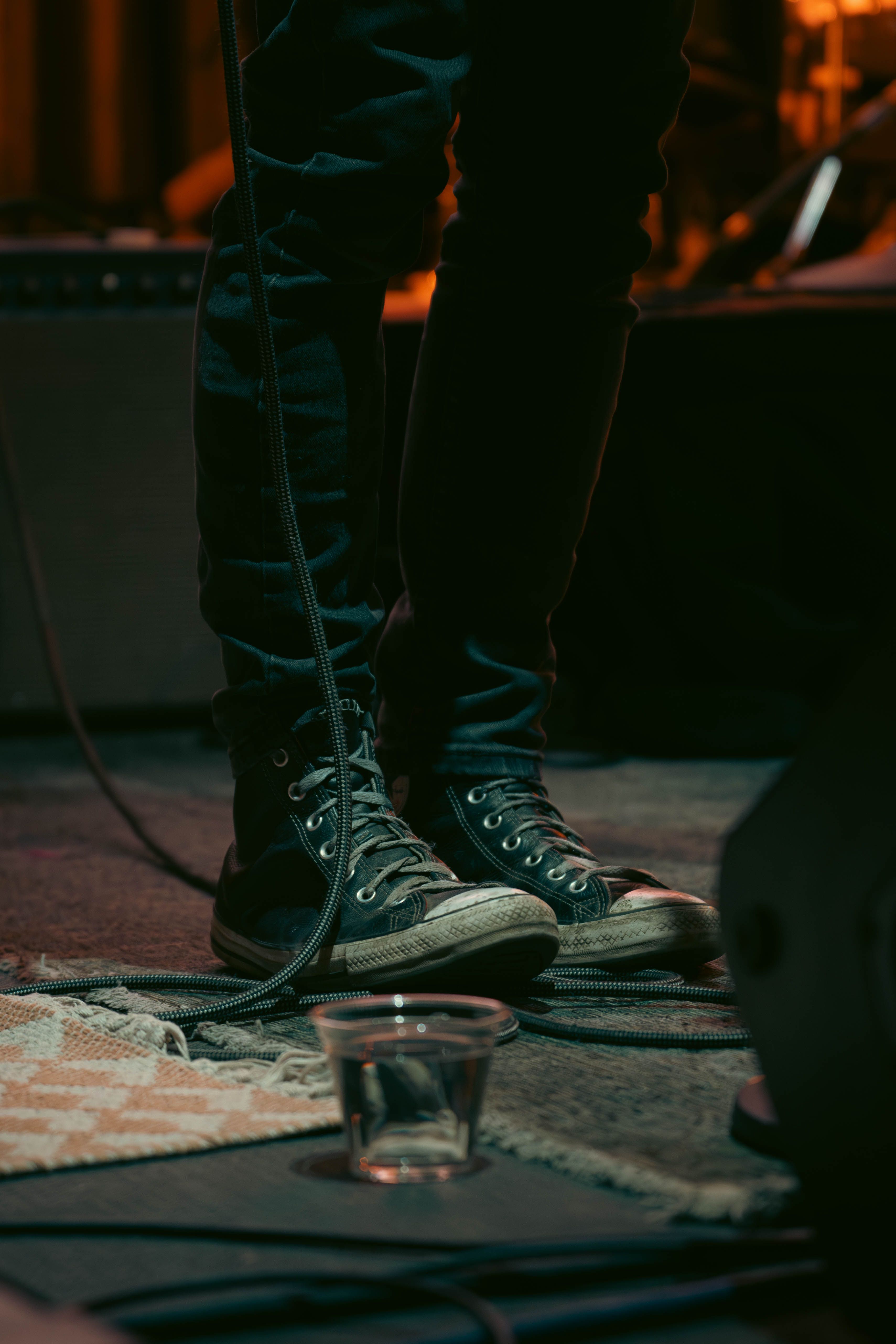 Close-up of a person wearing Converse sneakers on a dimly lit stage with microphone cable.