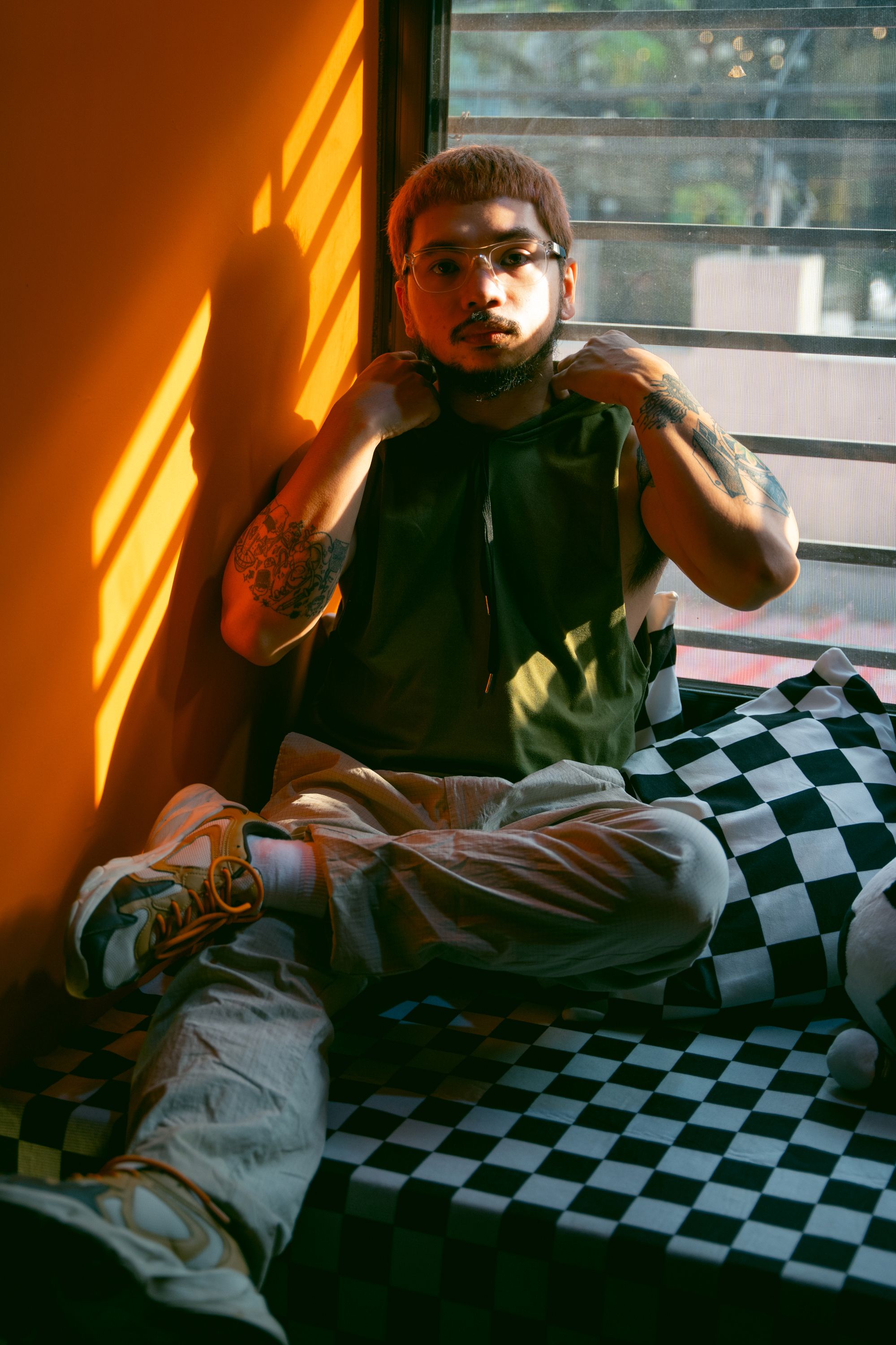 A man with tattoos relaxes on a checkered cushion by a sunlit window indoors.