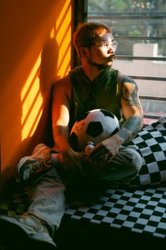 Man sitting by window with a soccer ball, bathed in warm evening light.