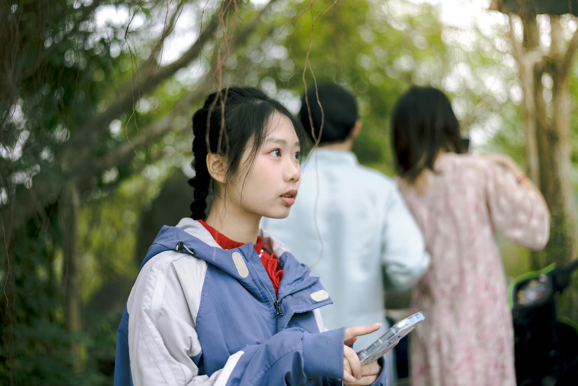 A young woman in a blue jacket holding a tablet surrounded by greenery outdoors.