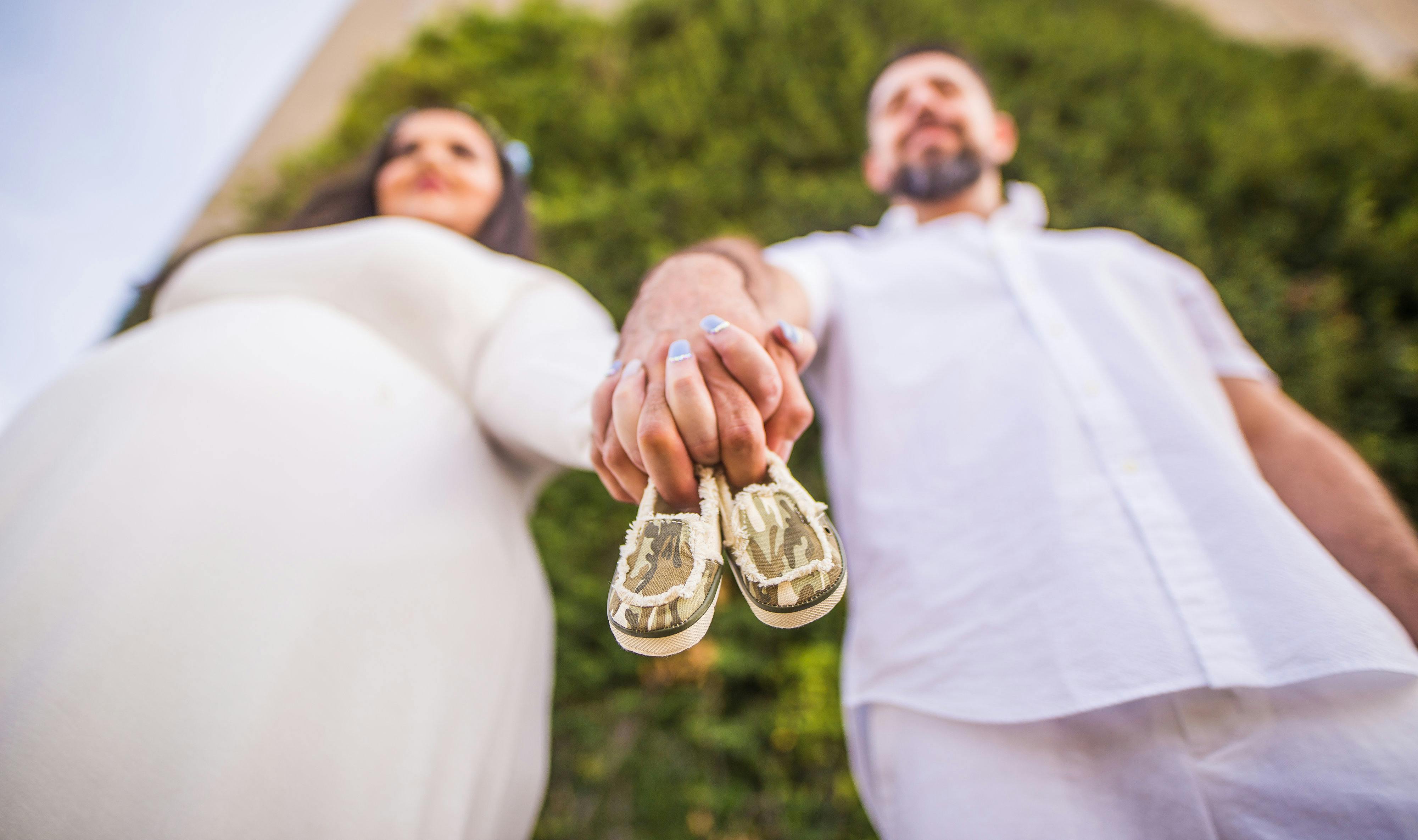 low angle shot of man and a pregnant woman holding hands with baby shoes between their fingers