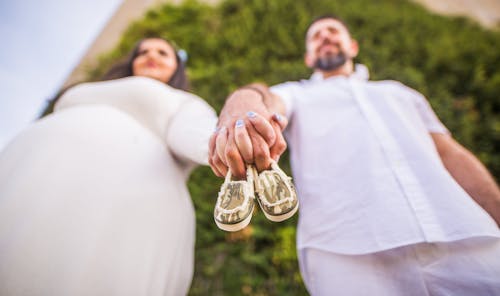Low Angle Shot Of Man and A Pregnant Woman Holding Hands With Baby Shoes Between Their Fingers