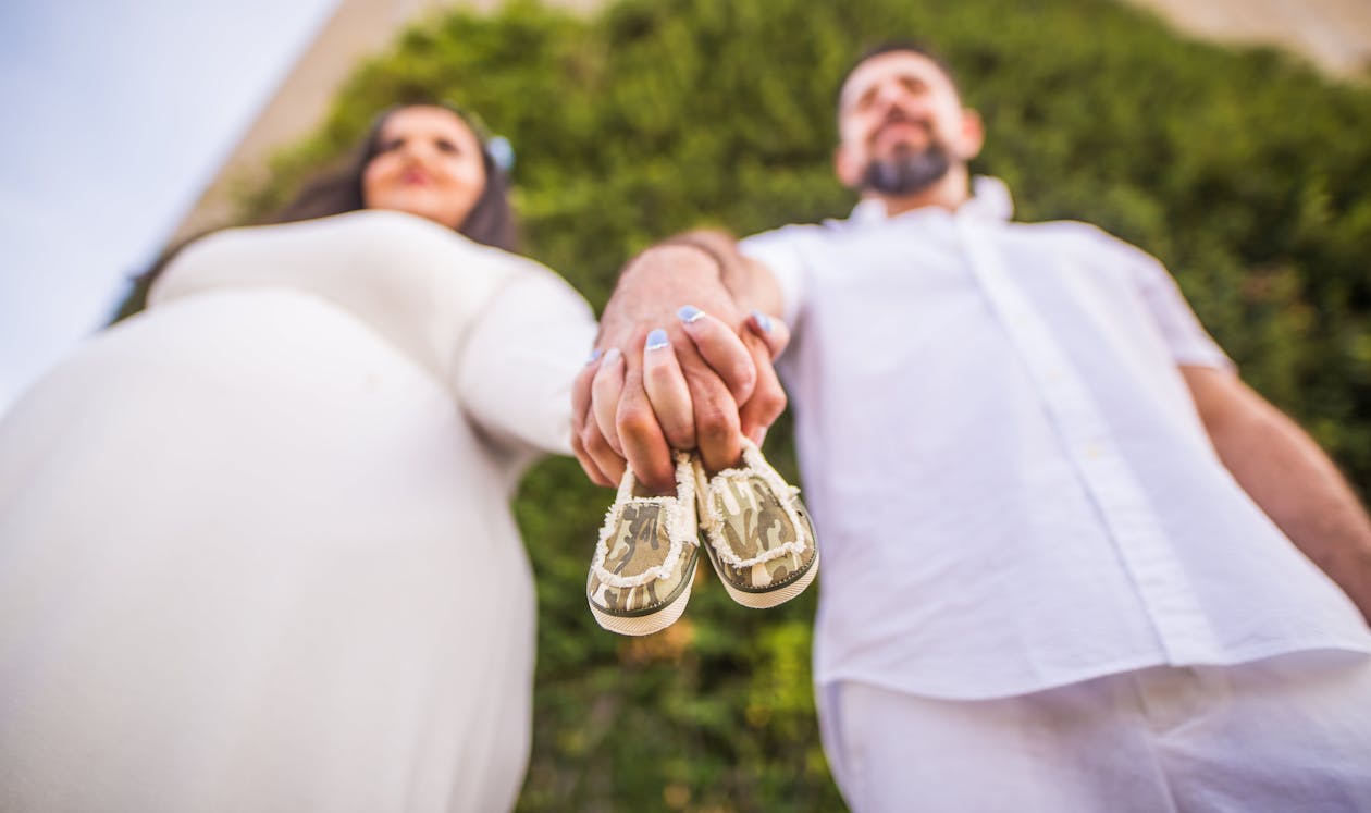 Low Angle Shot Of Man and A Pregnant Woman Holding Hands With Baby Shoes Between Their Fingers