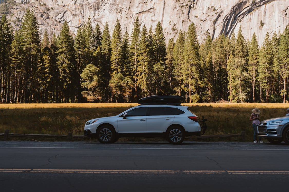 Vehicles Parked On The Side Of The Road Lined With Trees On The Foot Of A Rocky Mountain