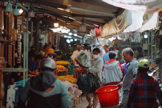 Busy street market scene with people buying and selling flowers and goods.