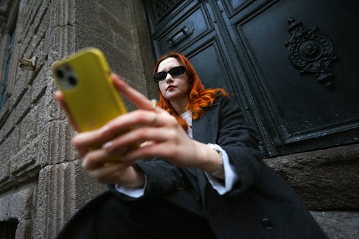 Fashionable woman with red hair taking a selfie in urban setting, wearing sunglasses.
