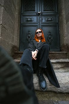 Fashionable woman in dark attire and sunglasses on stone steps before an ornate door.