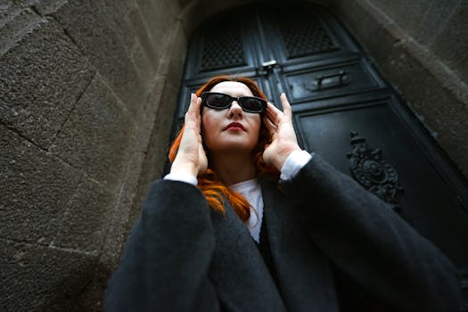 Confident woman in black coat and sunglasses posing by a gothic doorway.