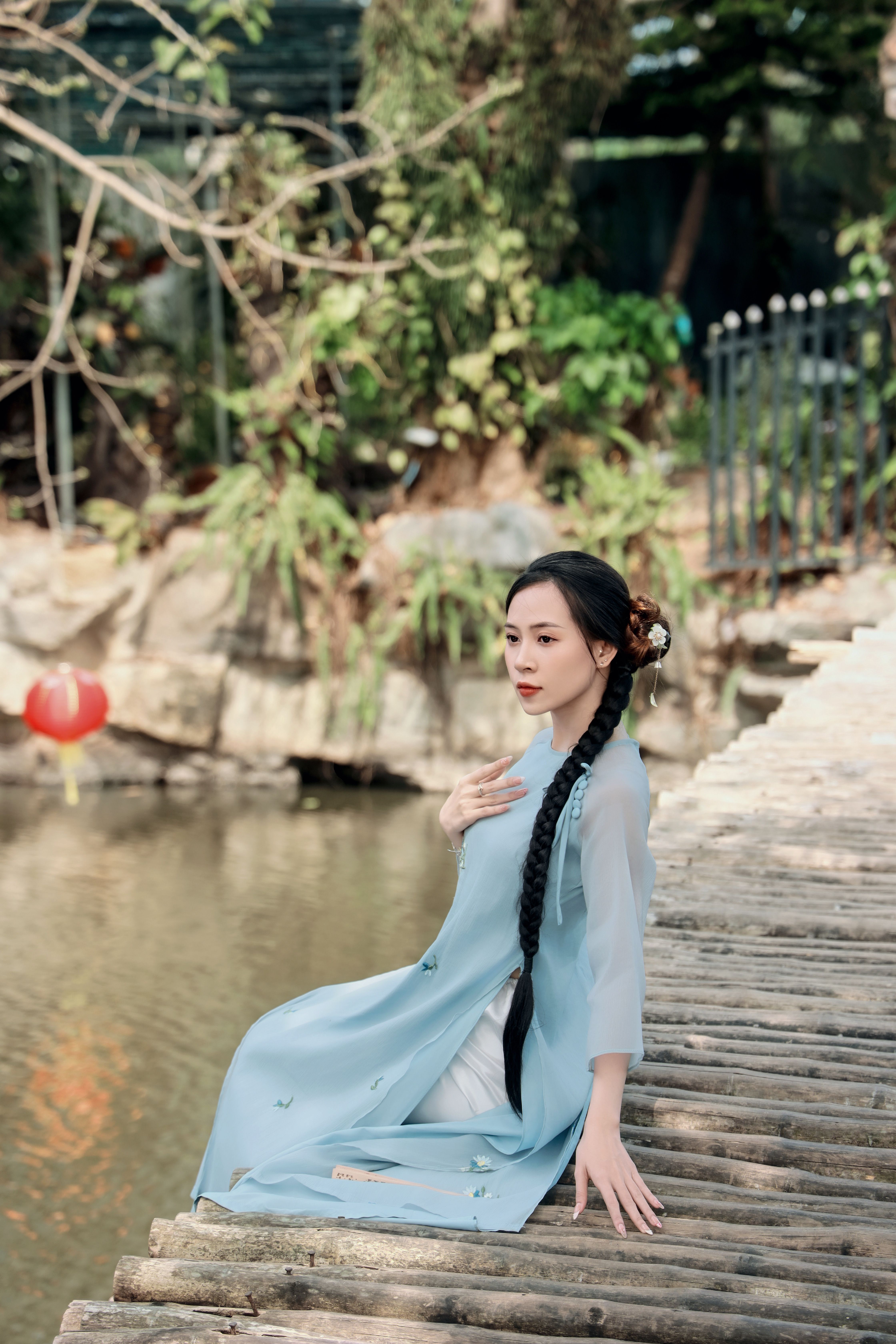 A woman in traditional attire sitting by a peaceful pond, surrounded by lush greenery.