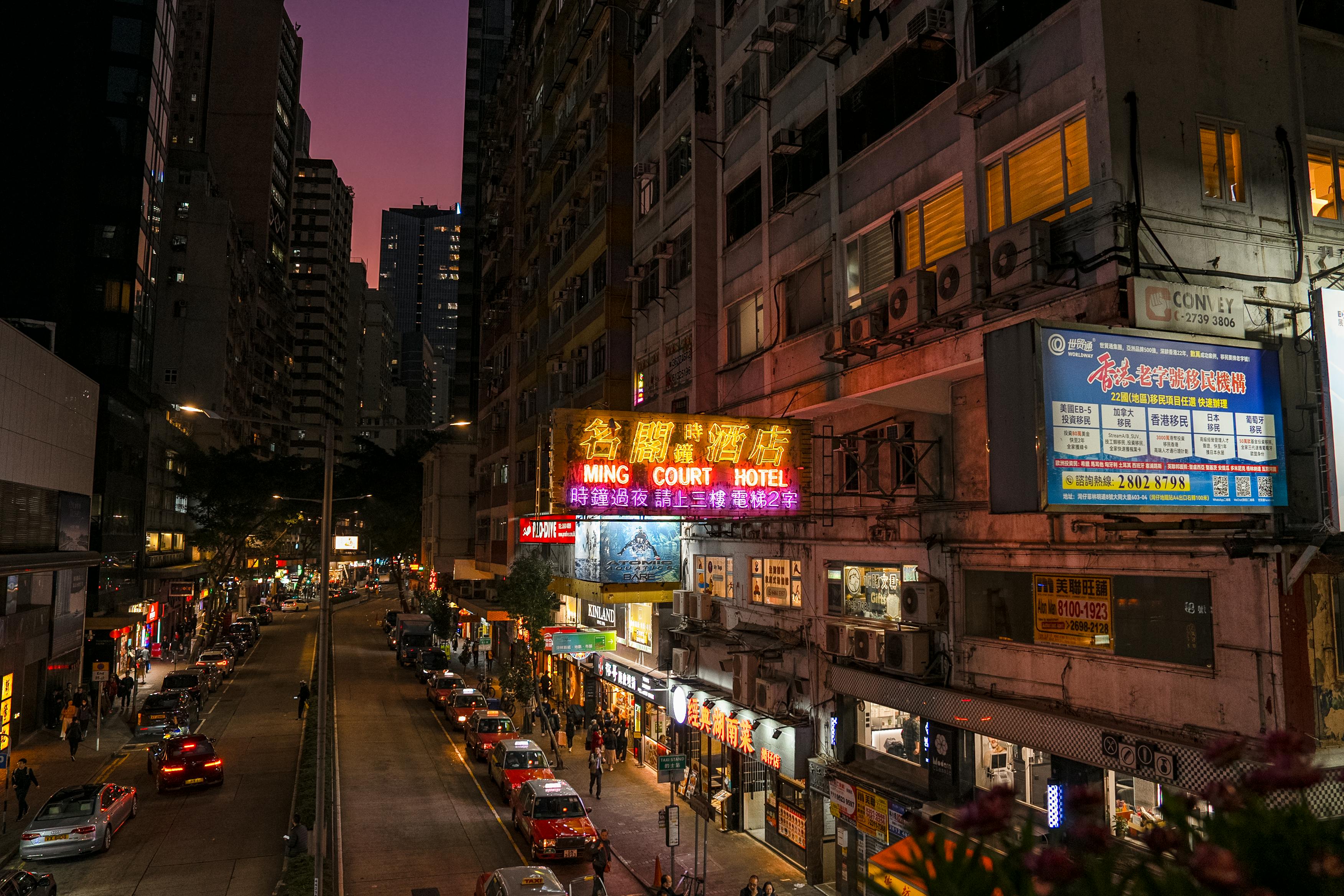 Bustling city street at dusk with colorful neon signs and urban architecture.