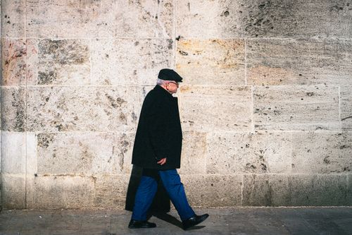 Free An elderly man in a coat walks alongside a large stone wall outdoors. Stock Photo