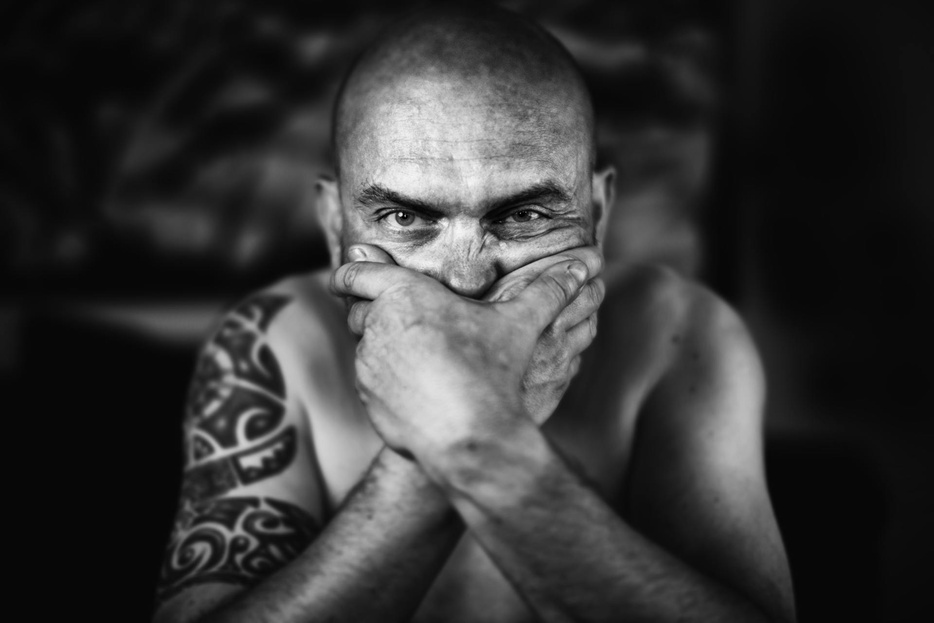 A powerful black and white portrait of a bald man with tattoos, covering his mouth with his hands.