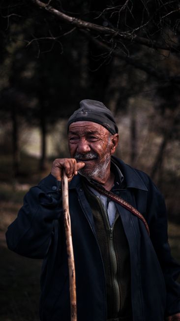 A senior man with a walking stick stands outdoors in a forested area.