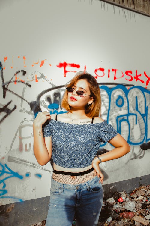 Женщина в солнцезащитных очках и блузке с открытыми плечами стоит у стены с граффити и мусором