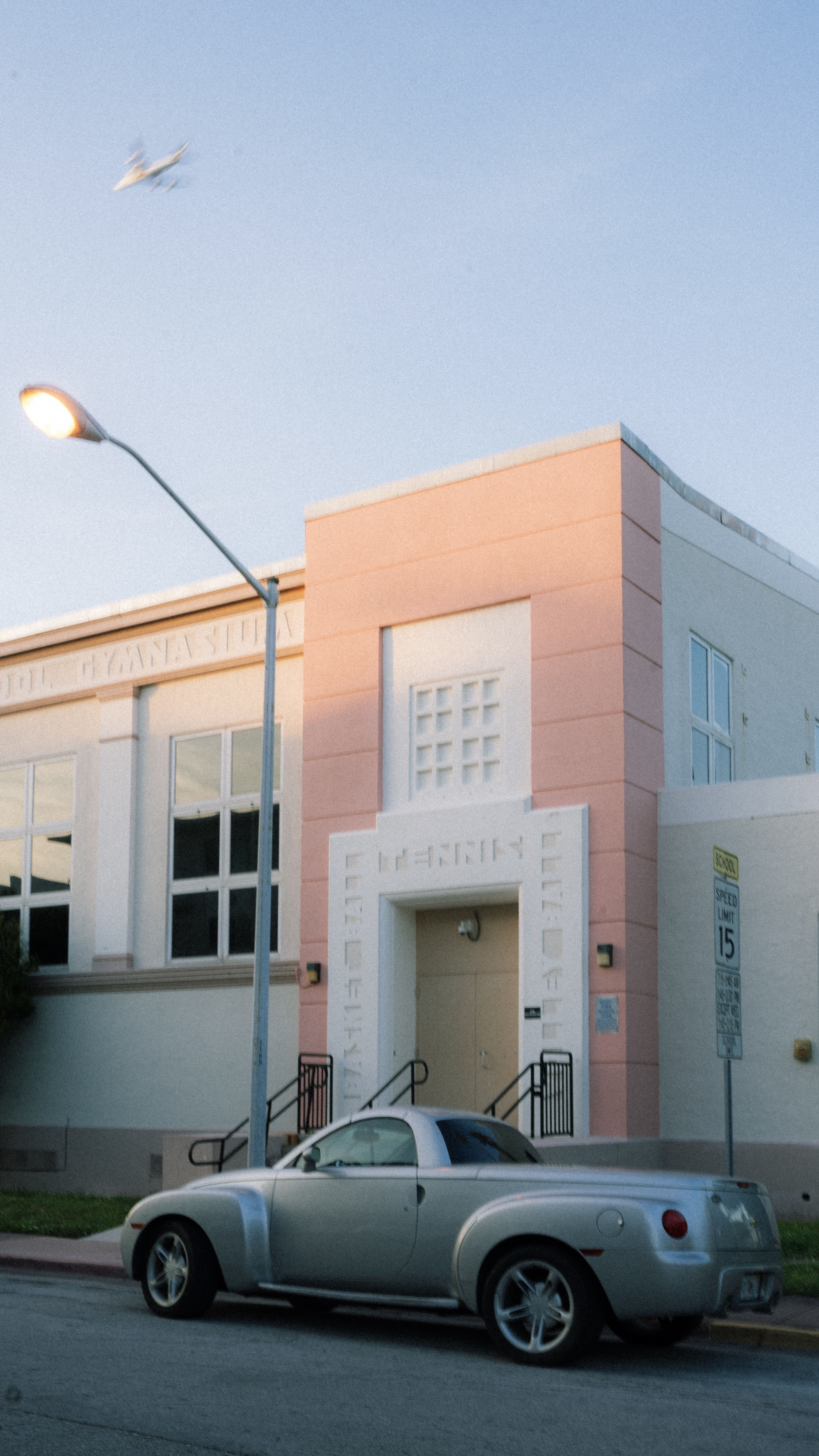 A vintage-style car parked in front of an Art Deco building with a plane flying overhead at sunset.