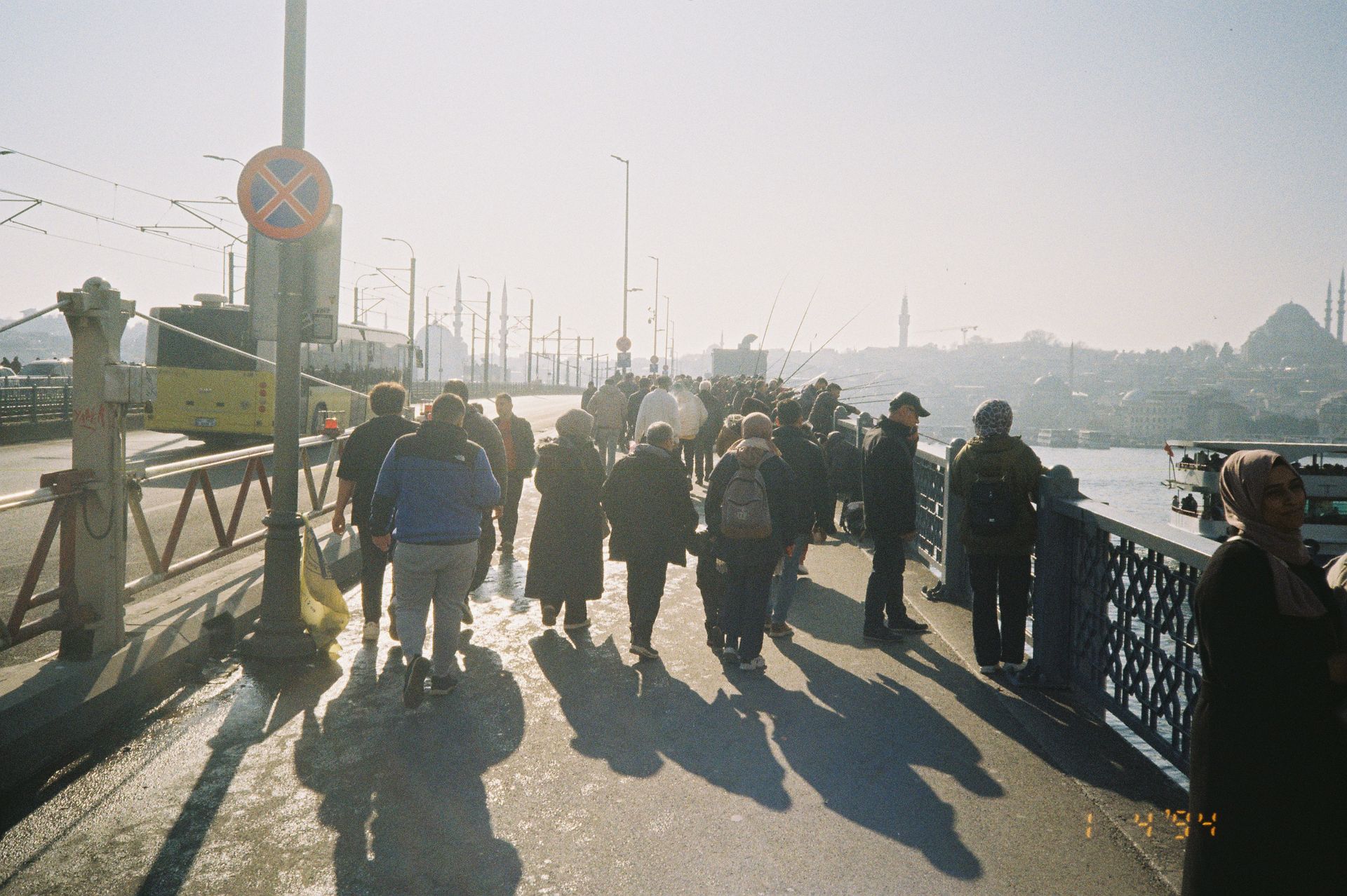 A crowd of people walks across a city bridge under bright sunlight, with a distant cityscape visible.
