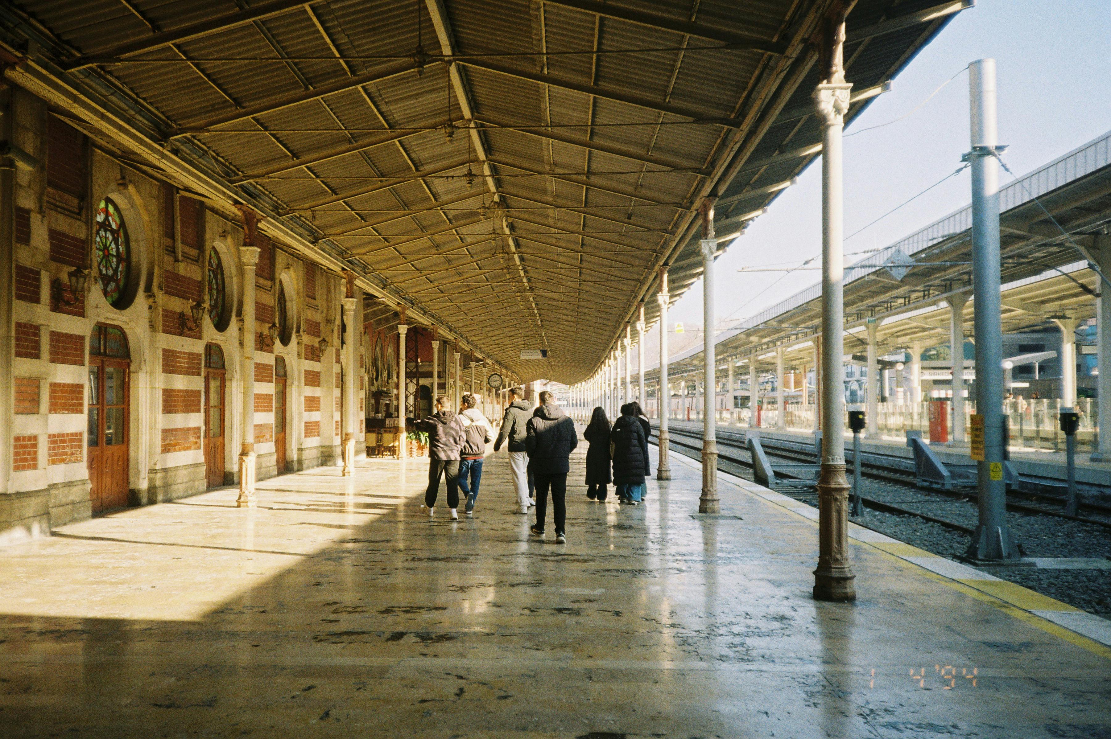 A group of people walking on a sunlit historic railway platform with arches.