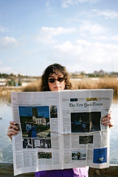 Woman reading a newspaper outdoors by a tranquil lake, sunny day.