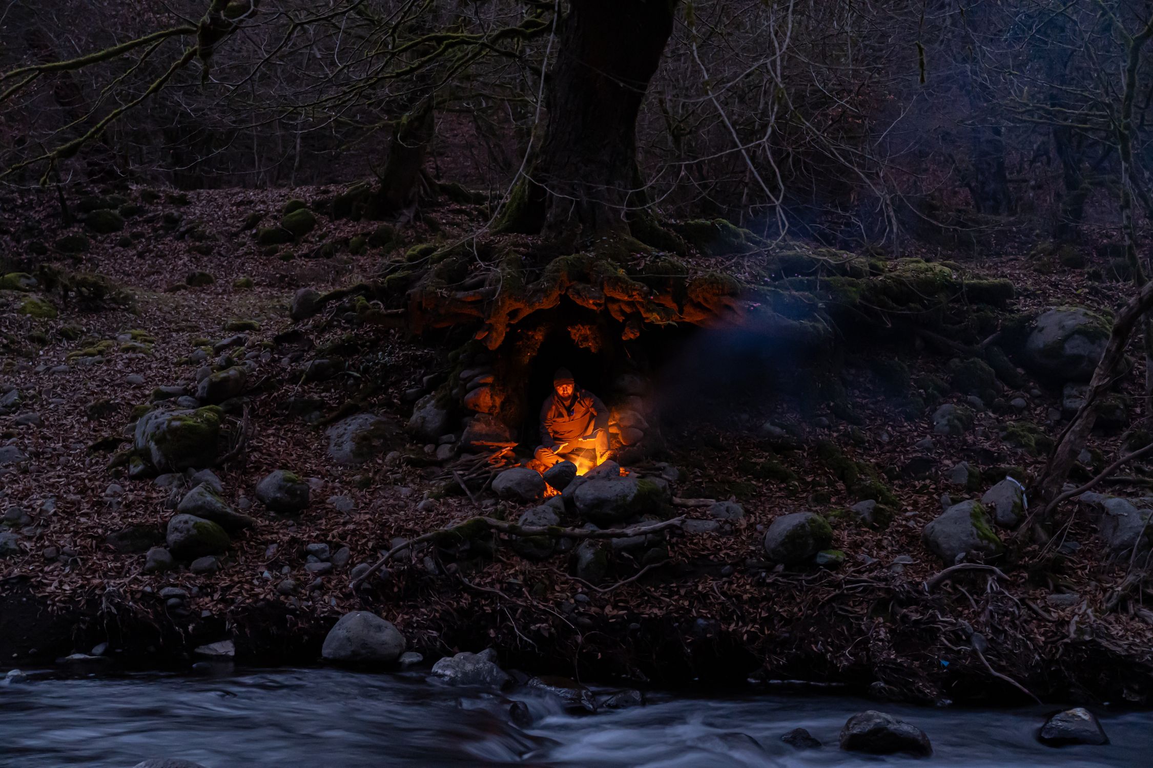 A lone camper sits by a cozy campfire in a cave surrounded by a dark forest.