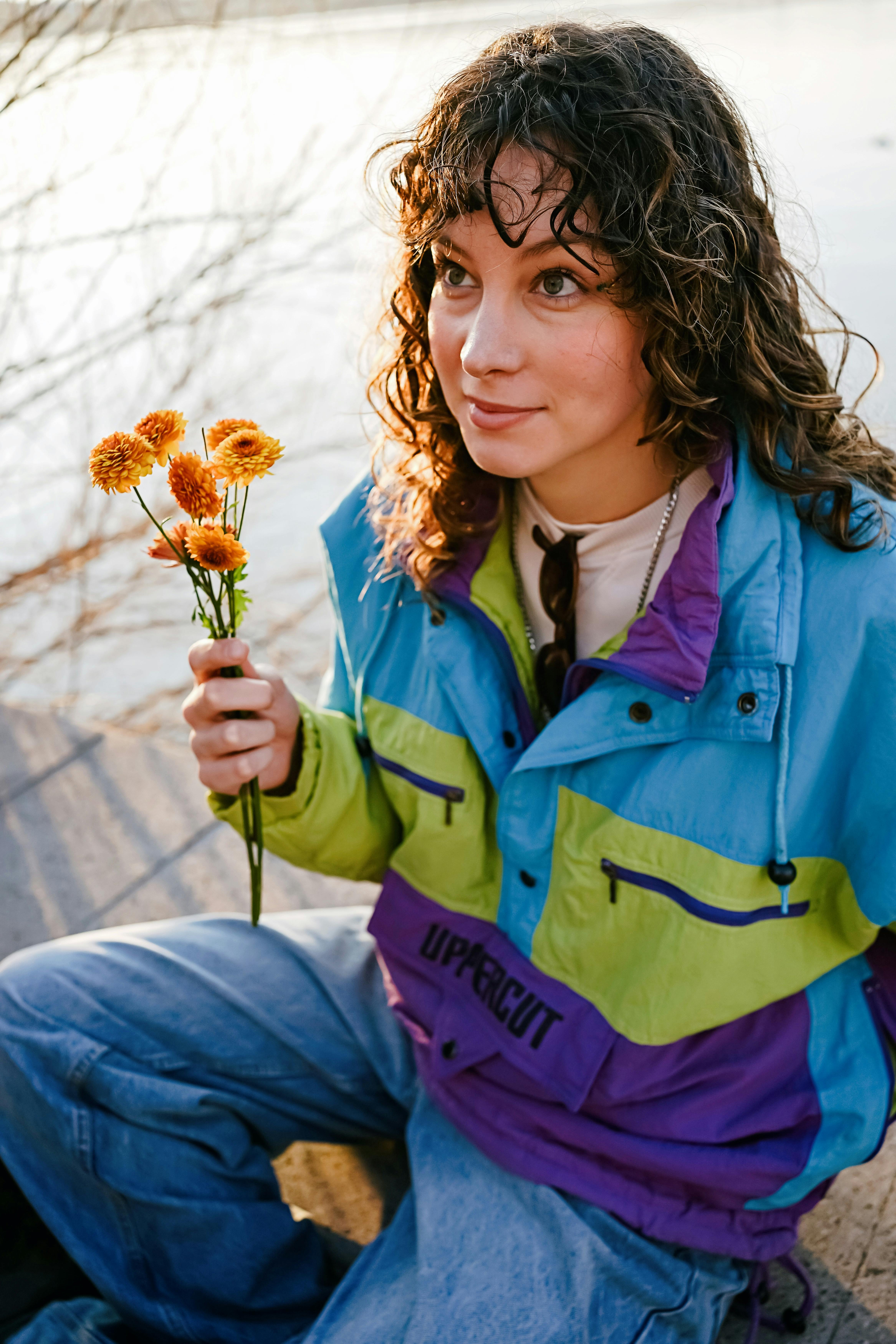 Smiling woman holding flowers, wearing a colorful jacket by a lake.
