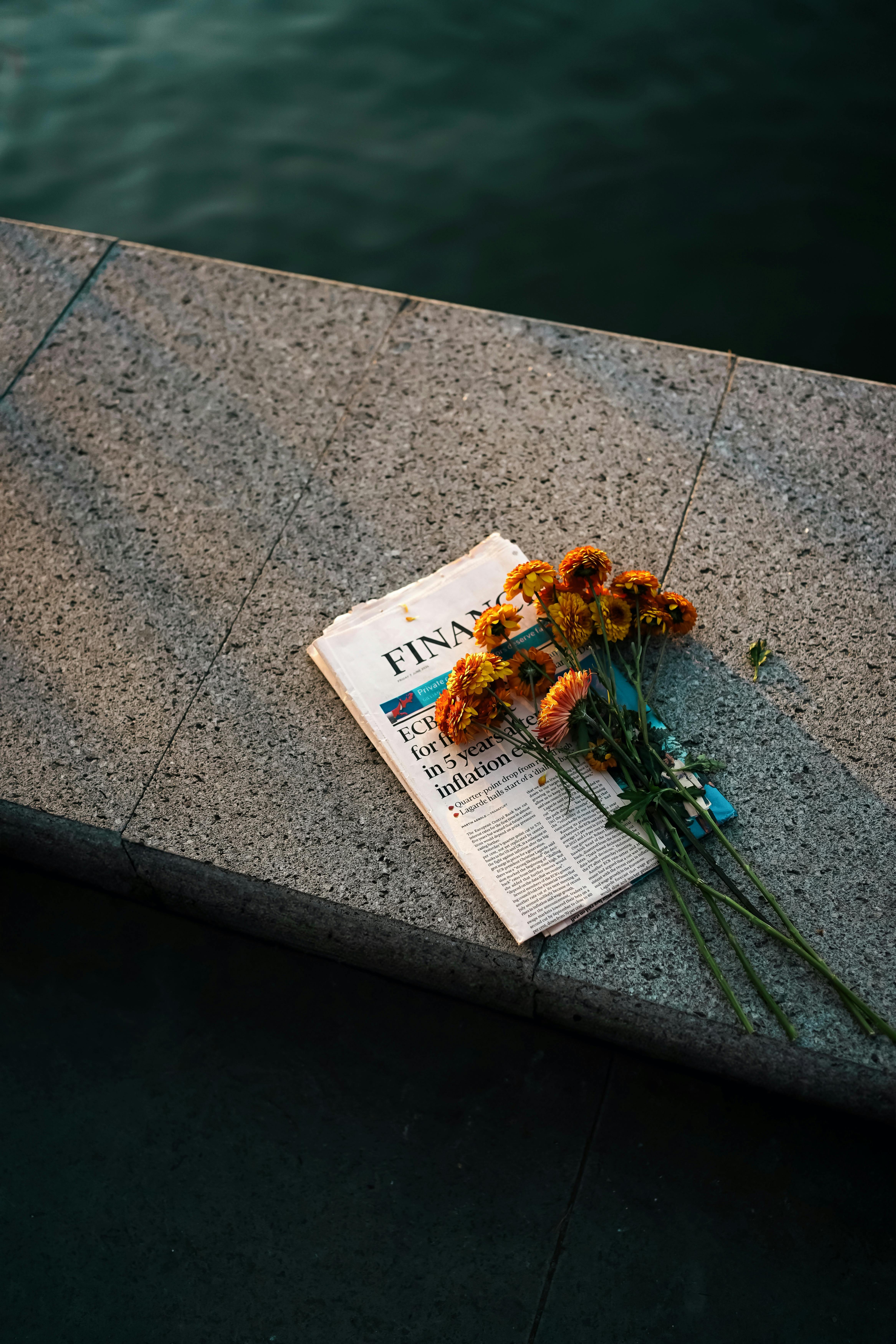 A newspaper and flowers rest on a stone seat beside the water at sunset, creating a serene moment.