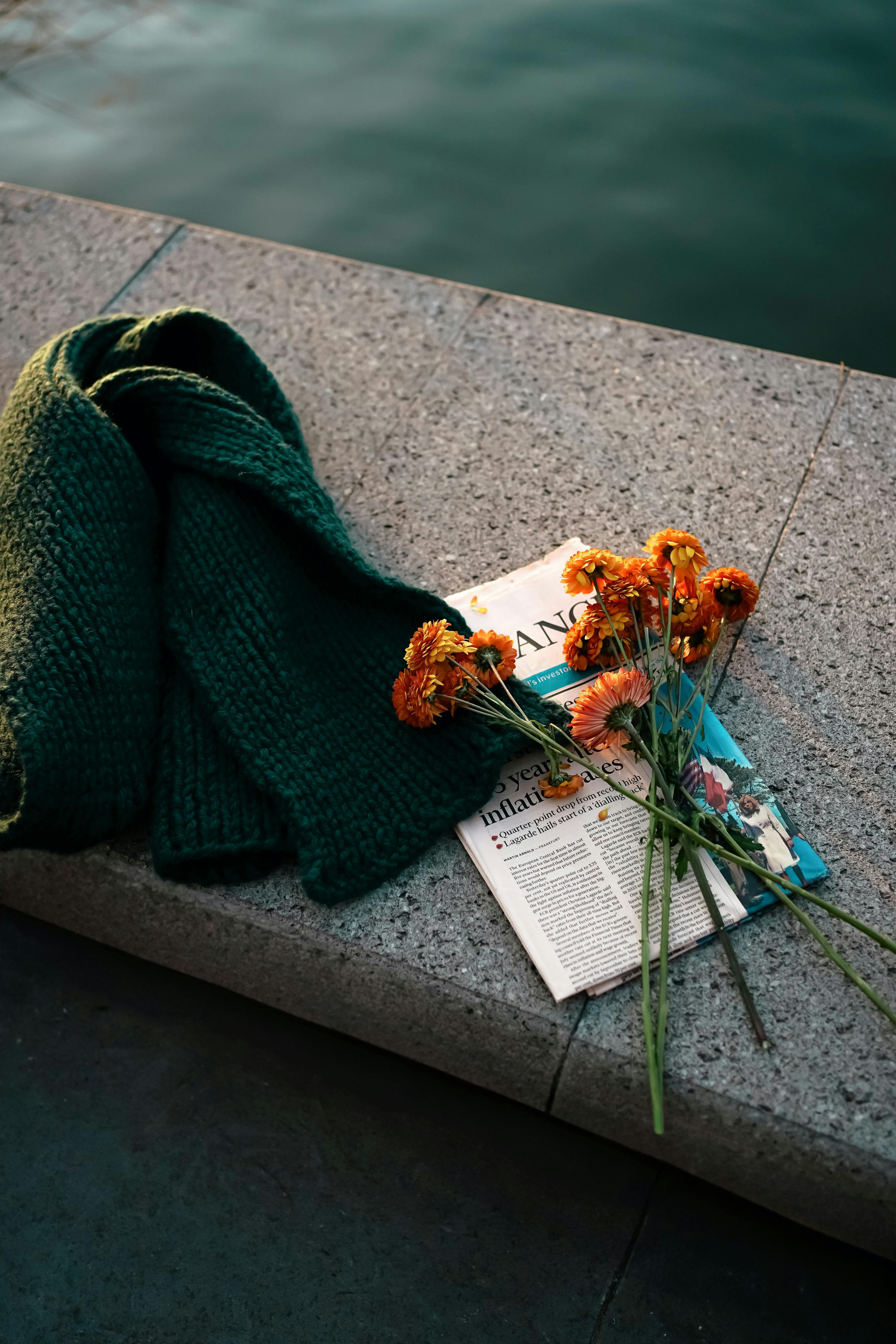 Green knit scarf and marigolds on stone bench with water background.