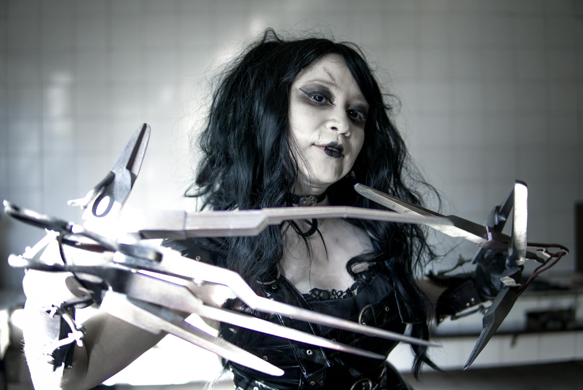 A woman in gothic cosplay with elaborate scissorhands in a dimly lit indoor setting.