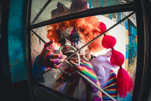 Free Vibrant horror clown peering through a broken window, showcasing eerie makeup and dramatic colors. Stock Photo