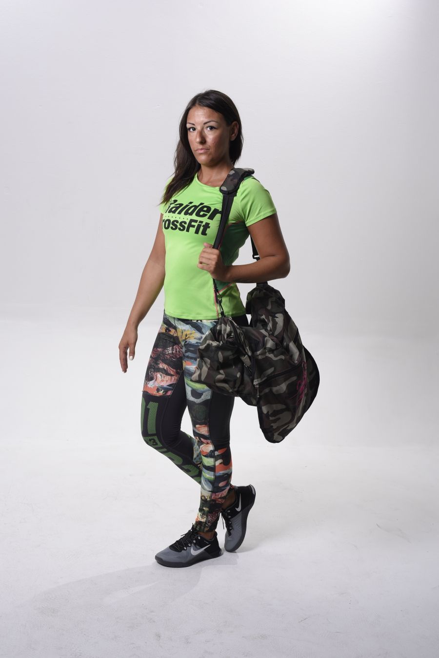 Athletic woman in vibrant gym clothes posing with a camo duffel bag against a clean background. Stock Photo