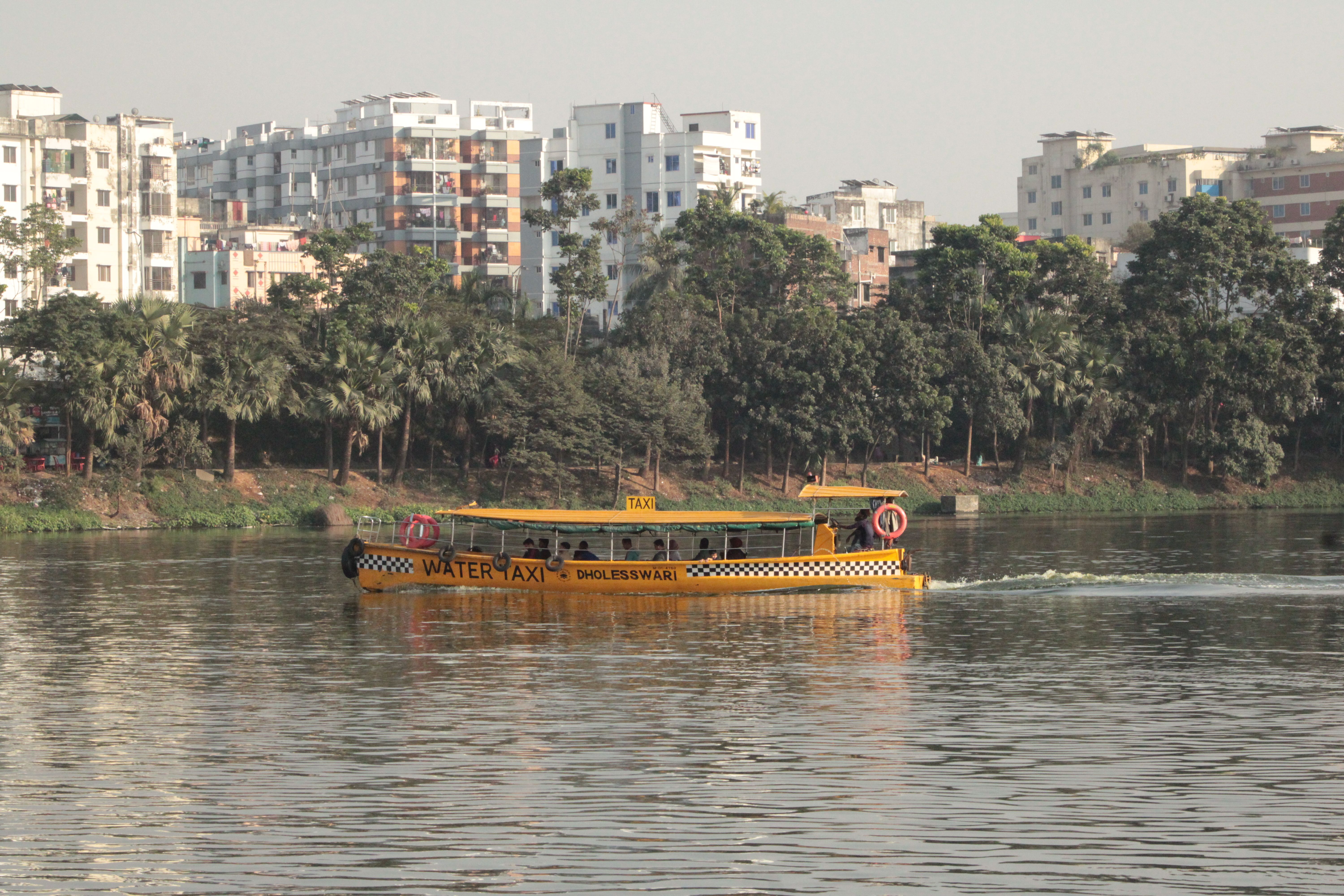 Yellow water taxi navigates Dhaleshwari River with city buildings in background.