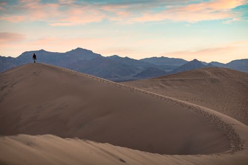 Free A solitary figure walks atop sand dunes at dusk with mountains in the background. Stock Photo