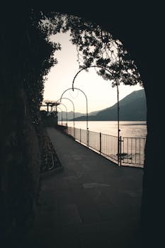 Tranquil lakeside pathway with arches and mountains at Lake Como, Italy at twilight.