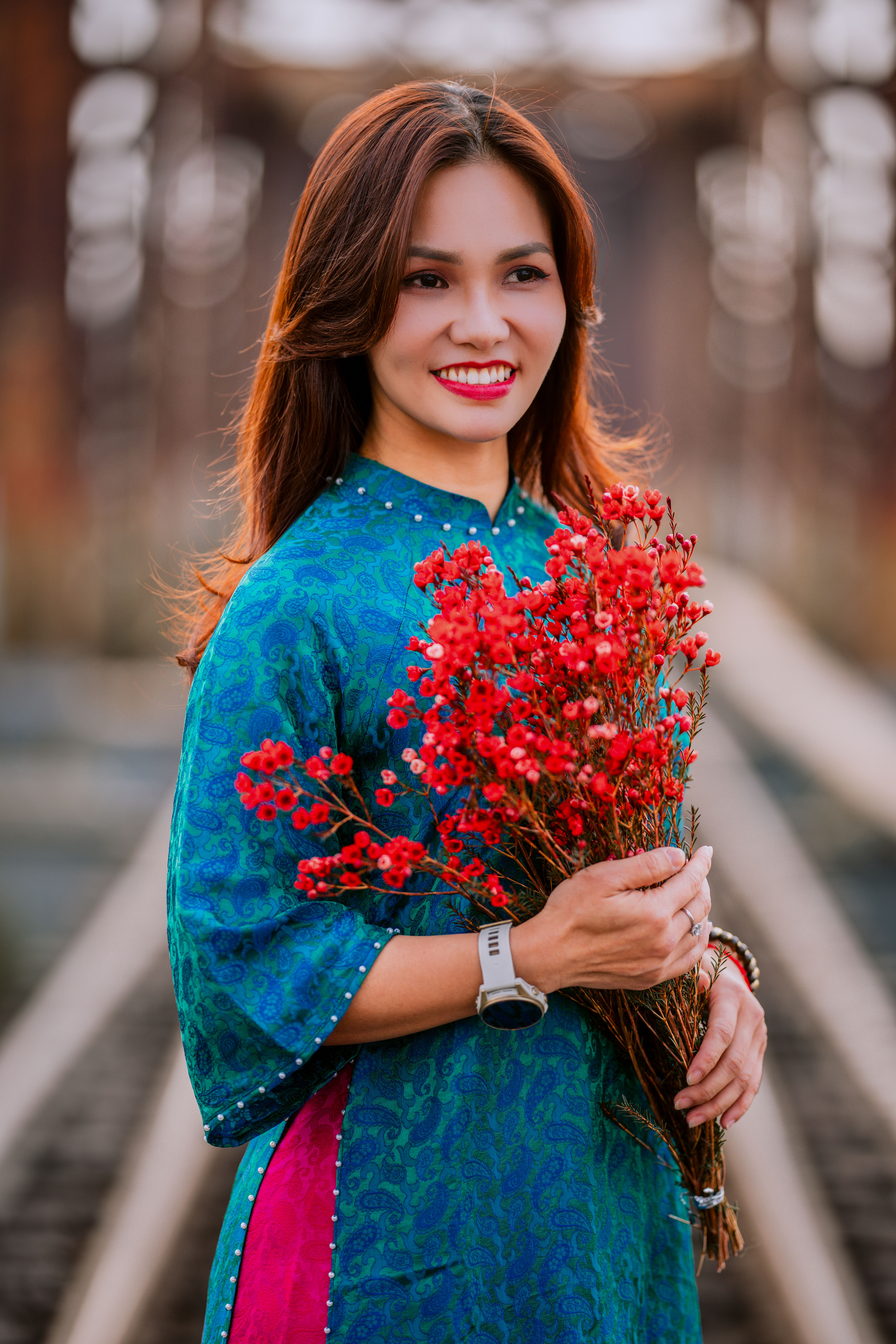 Woman in blue traditional dress holding red flowers, smiling on a railway track.