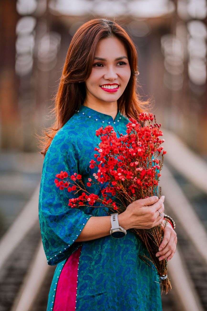 Woman in blue traditional dress holding red flowers, smiling on a railway track.