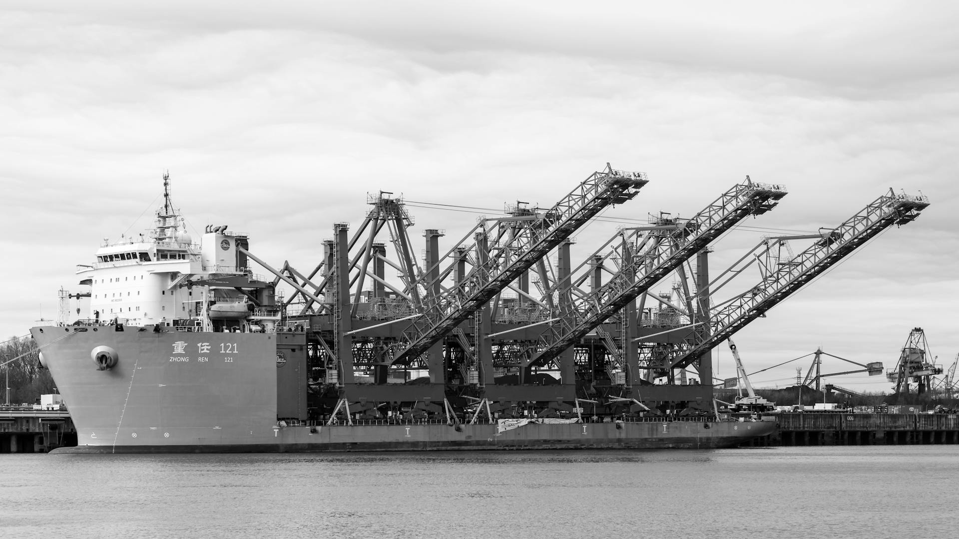 Black and white image of a container ship docked at Hamburg port under cloudy skies.