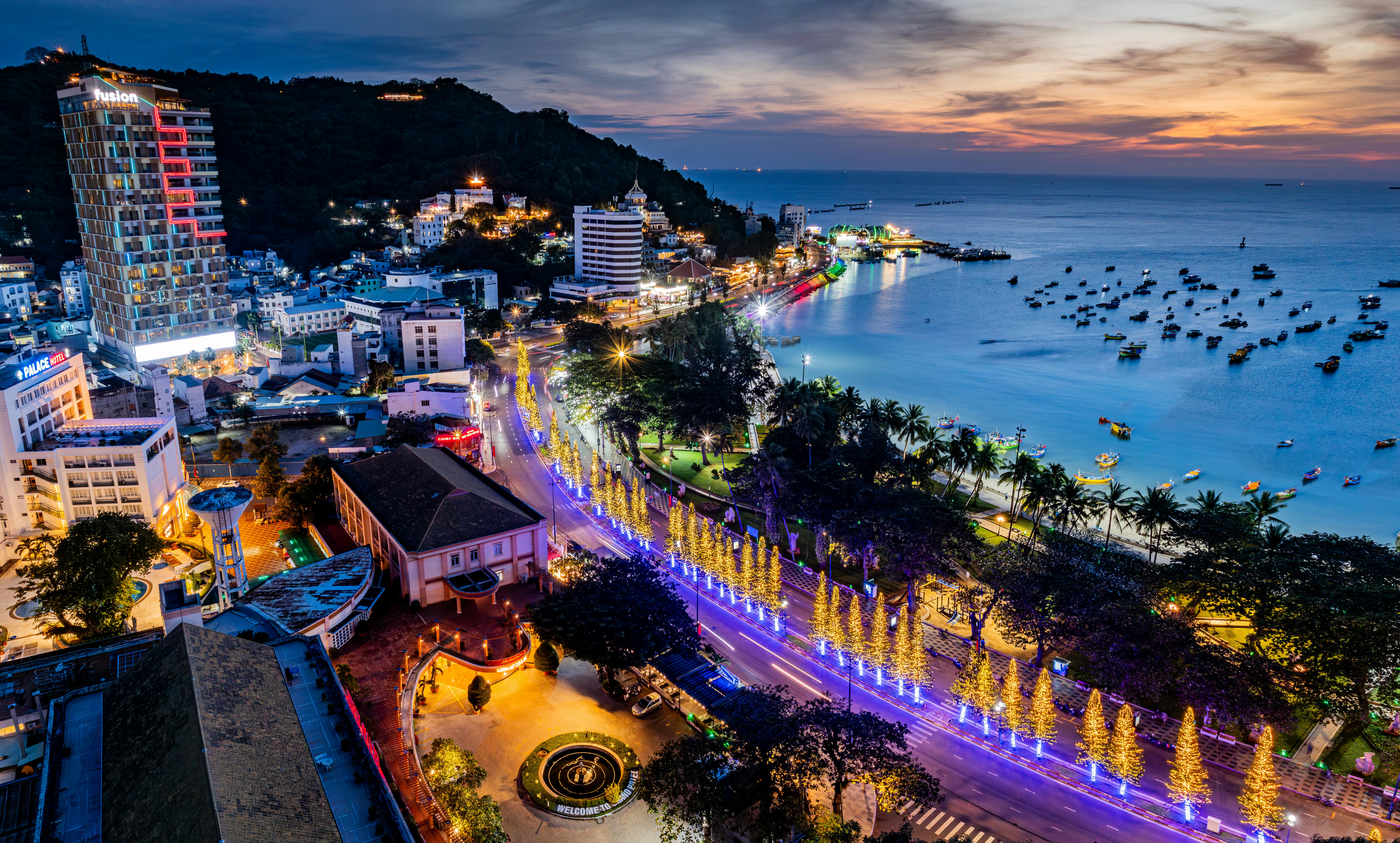 Stunning aerial view of a coastal cityscape at twilight, showcasing festive lights and a serene beach.