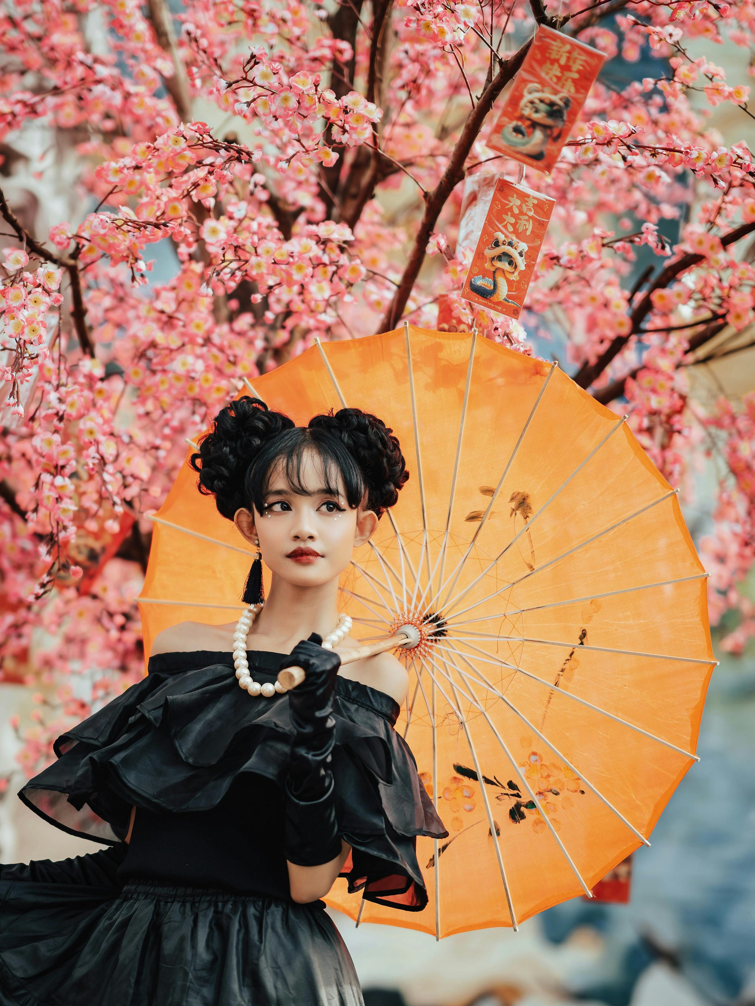 Woman in black dress with orange parasol stands by cherry blossom tree, evoking spring elegance.