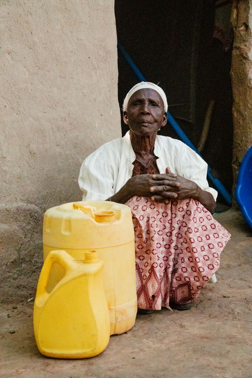Free An elderly woman sits outdoors beside yellow water containers in a rural setting, showcasing daily life. Stock Photo