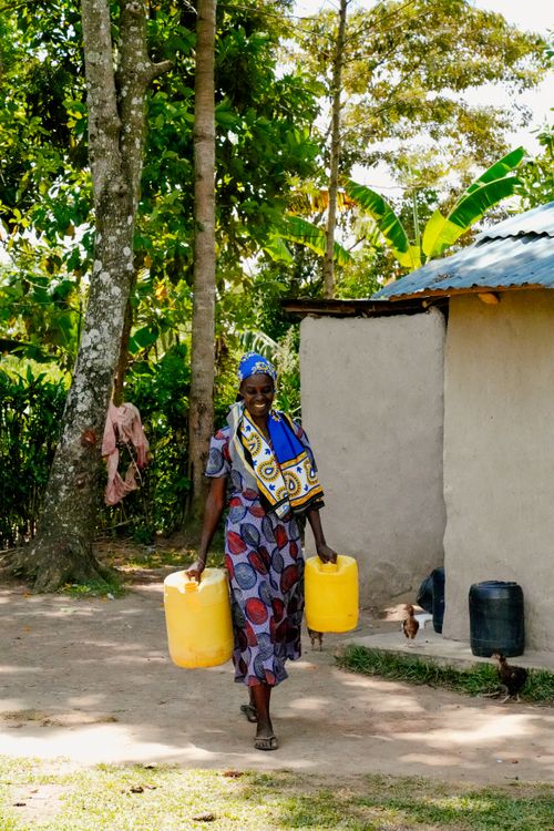 Free African woman carrying yellow plastic jugs of water next to a traditional house surrounded by lush greenery. Stock Photo