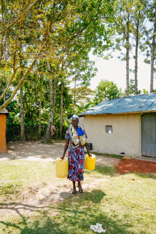 Free An African woman in traditional attire carrying yellow water jugs in a rural setting. Stock Photo
