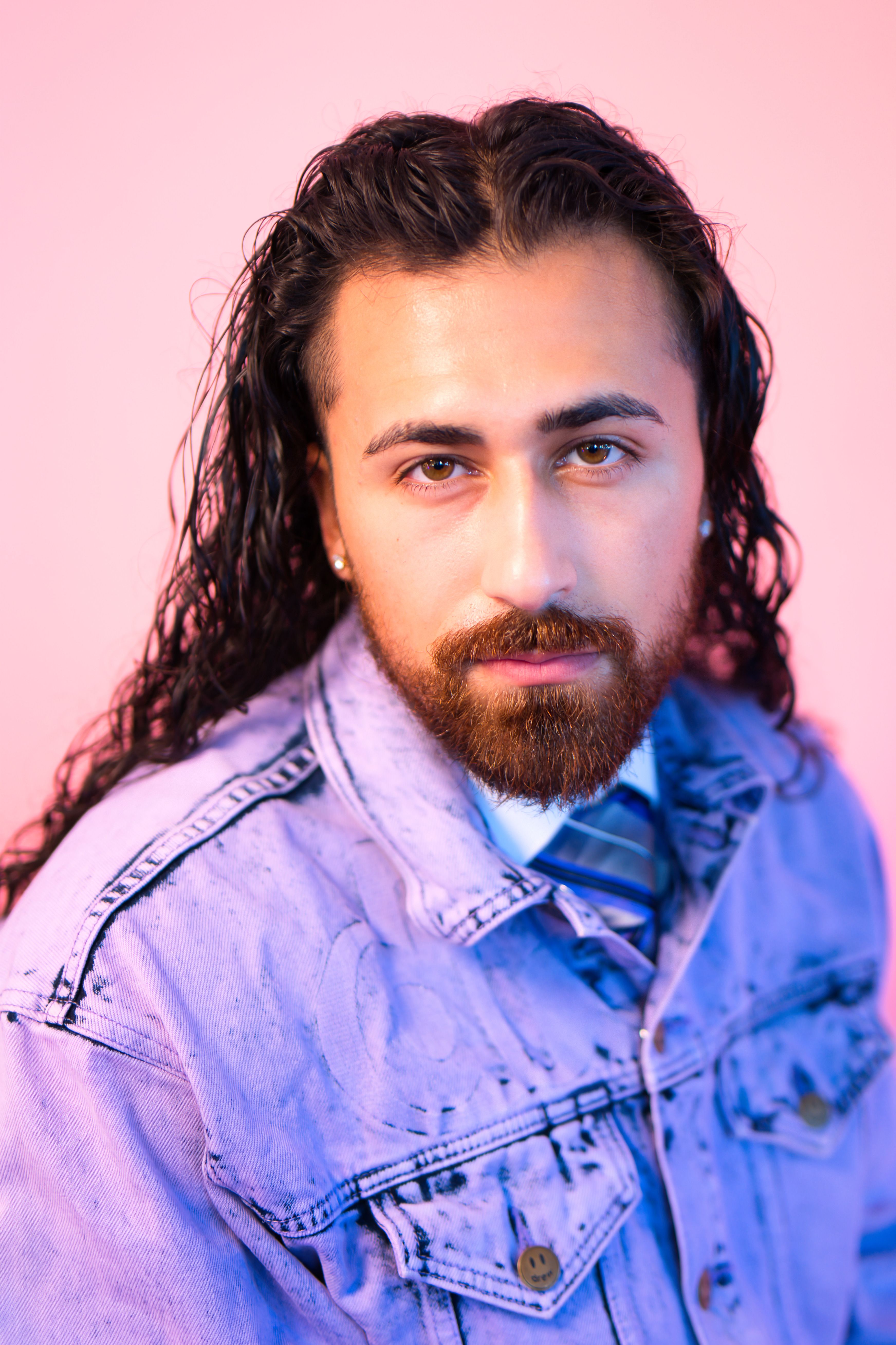 Stylish man with long hair and beard wearing denim jacket against pink background.