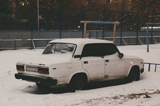 A vintage white car covered in snow parked in an urban setting during winter.
