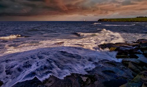 Free Powerful waves crash on rocky shore under a dramatic sunset sky with a distant lighthouse. Stock Photo