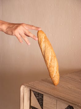 A hand balancing a baguette precariously on the edge of a wooden table indoors.