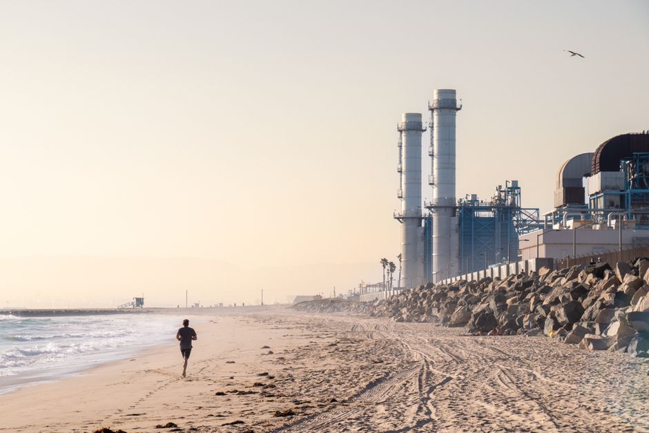 A person jogs along a sandy beach with an industrial plant nearby during sunrise.