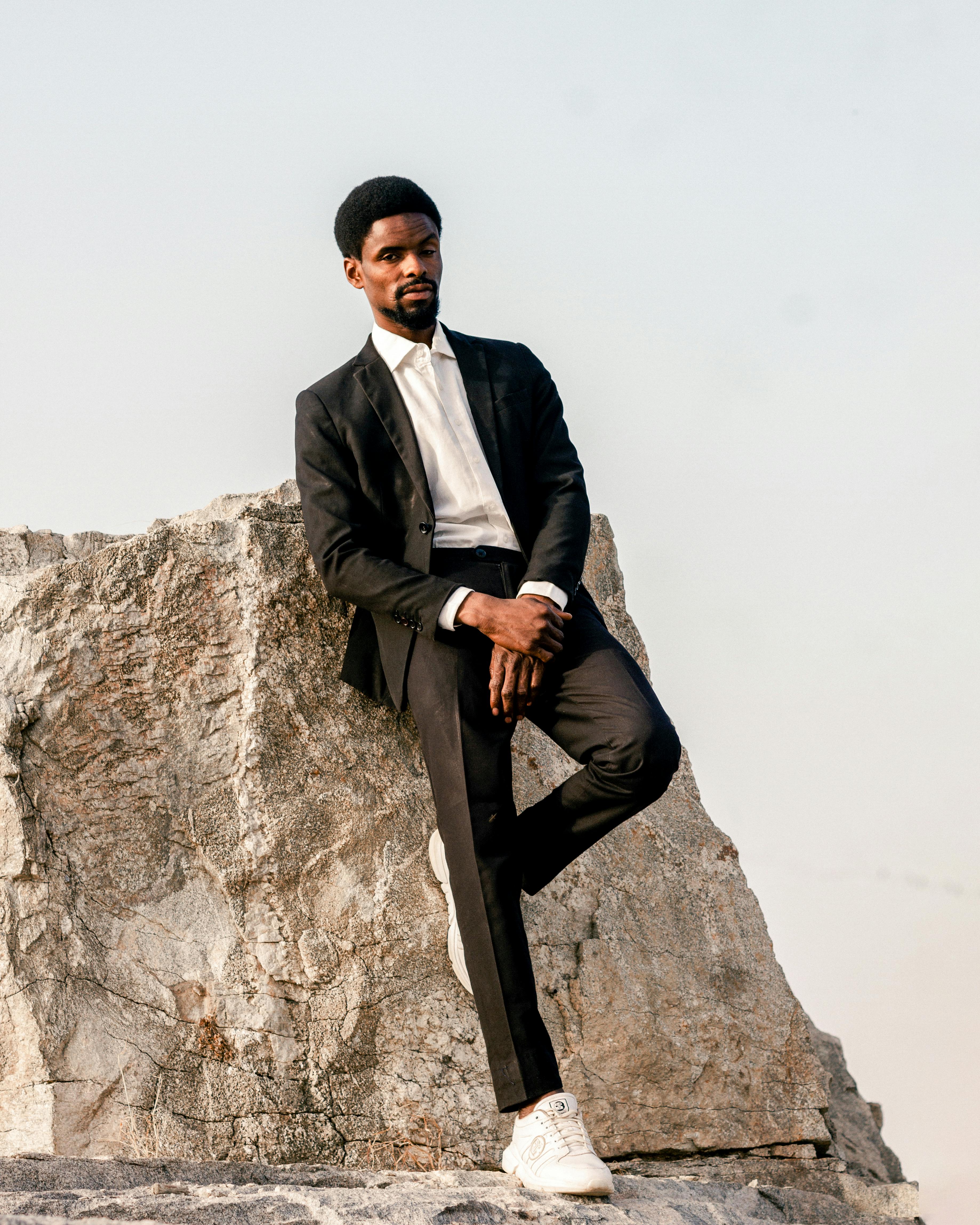 A fashionable man in a suit poses confidently on a large rock outdoors.