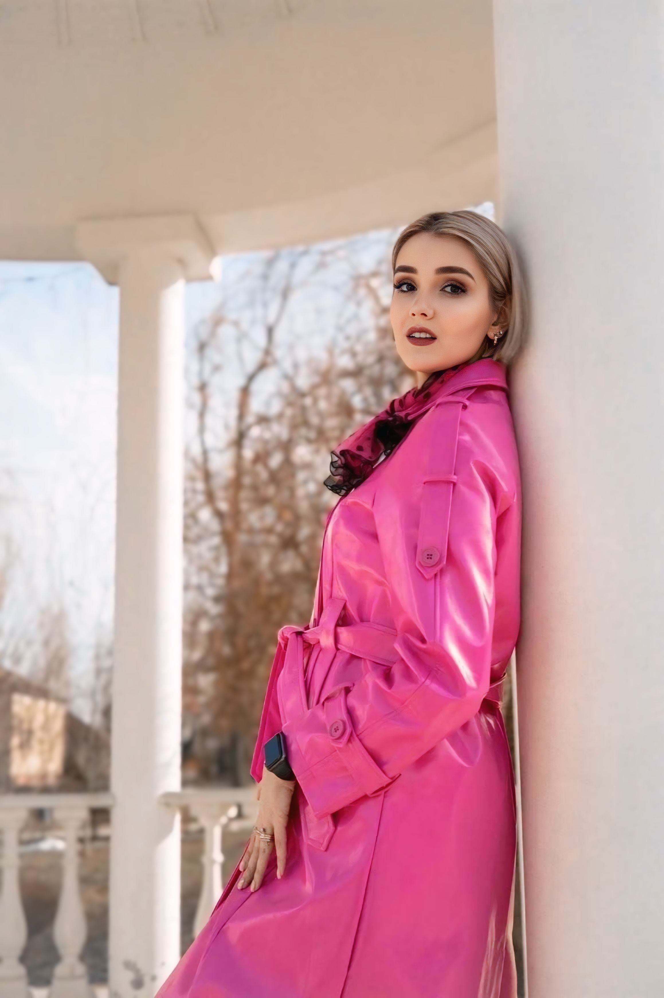 Stylish woman in a pink trench coat posing against a column outdoors.