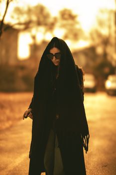 A woman wearing sunglasses and a dark shawl walks on a sunlit street during golden hour.
