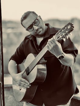 A man wearing sunglasses playing an acoustic guitar with a blurred outdoor background.